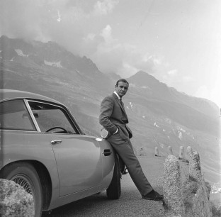 SEAN CONNERY relaxes on the bumper of his Aston Martin DB5 during the filming of location scenes for 'Goldfinger' in the Swiss Alps.